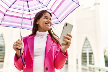 Young latin woman using smartphone holding umbrella at street