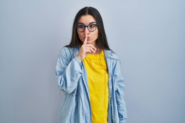 Mavi arka planda dikilen genç İspanyol kadın, parmakları dudaklarında sessiz olmak istiyor. sessizlik ve gizli konsept. 