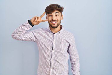 Arab man with beard standing over blue background doing peace symbol with fingers over face, smiling cheerful showing victory 