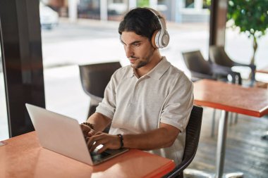 Young hispanic man using laptop and headphones at coffee shop terrace