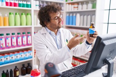 Young hispanic man pharmacist holding medication bottle speaking at pharmacy