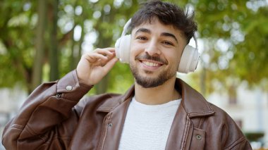 Genç Arap adam parkta müzik dinlerken kendinden emin bir şekilde gülümsüyor.