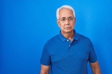 Mavi arka planda duran gri saçlı, kuşkucu ve gergin orta yaşlı bir adam sorun yüzünden surat asıyor. negatif kişi. 