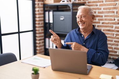Gri saçlı yaşlı adam ofiste bilgisayar laptopuyla çalışıyor gülümsüyor ve kameraya bakıyor iki eliyle ve parmaklarıyla onu işaret ediyor.. 