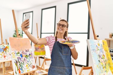 Gülümseyen genç bir kadın sanat stüdyosundaki akıllı telefondan özçekim yapıyor.