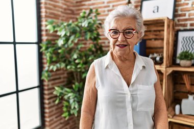 Yaşlı, gri saçlı, gülümseyen bir kadın. Evde kendine güveniyor.