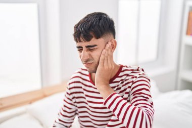 Young hispanic man suffering for ear pain sitting on bed at bedroom