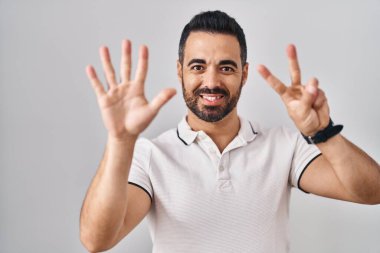 Beyaz arka plan üzerine günlük giysiler giyen, sakallı genç İspanyol adam yedinci parmaklarını gösterip gülümserken kendinden emin ve mutlu görünüyor.. 