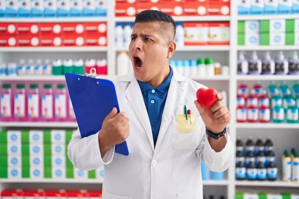 Young Hispanic Man Working Pharmacy Drugstore Holding Heart Angry Mad — Stock Photo, Image