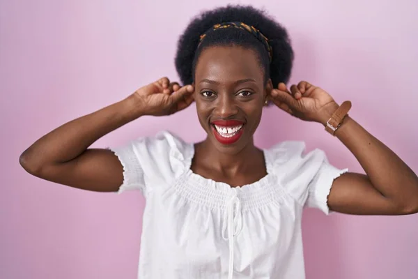 头发卷曲的非洲女人站在粉红的背景上 微笑着用手指拉耳朵 滑稽的手势 试镜问题 — 图库照片