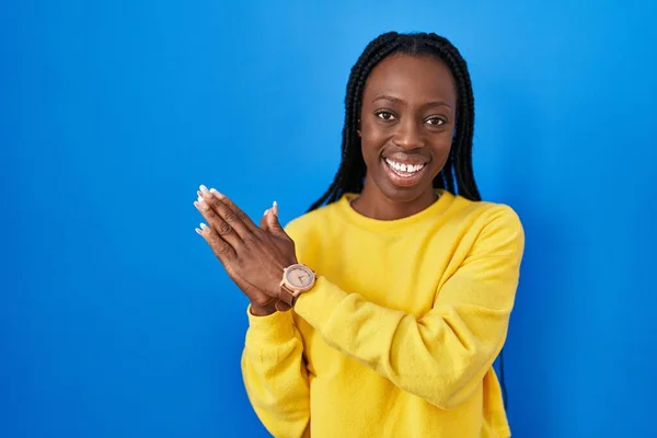 Beautiful black woman standing over blue background clapping and applauding happy and joyful, smiling proud hands together