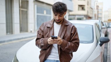 Genç Arap adam akıllı telefon kullanıyor. Sokakta arabaya yaslanıyor.