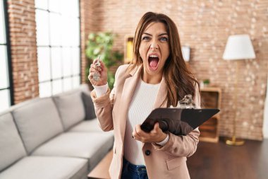 Young brunette woman working as real state agent holding keys of new home angry and mad screaming frustrated and furious, shouting with anger. rage and aggressive concept. 