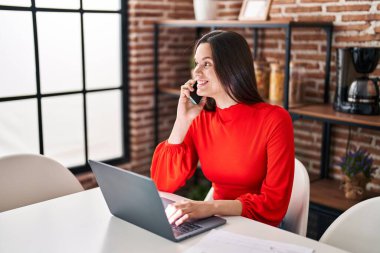 Young beautiful hispanic woman using laptop talking on smartphone at home