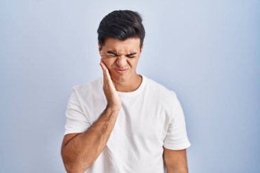 Mavi arka planda dikilen İspanyol bir adam diş ağrısı ya da diş hastalığından dolayı acı dolu bir ifadeyle ağıza dokunuyor. dişçi 