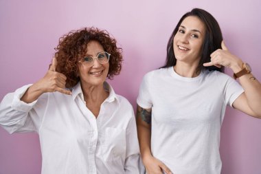 İspanyol anne ve kızı pembe arka planda rahat beyaz tişört giyip telefonla konuşurken gülümsüyorlar. iletişim kavramları. 