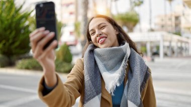 Genç ve güzel İspanyol kadın gülümsüyor. Kendine güveni tam. Sokakta akıllı telefonun yanında selfie çekiyor.