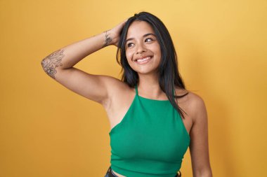 Sarı arka planda duran esmer kadın gülümsüyor. Saçlarına dokunuyor, el hareketi yapıyor, çekici ve şık duruyor. 