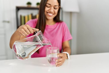 Genç Latin kadın gülümsüyor. Kendine güveni tam. Eve su dolduruyor.