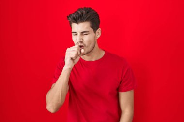 Kırmızı arka planda duran İspanyol genç adam kendini iyi hissetmiyor ve soğuk algınlığı veya bronşitin belirtisi olarak öksürüyor. sağlık hizmeti kavramı. 