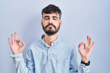 Mavi arka planda duran sakallı genç İspanyol adam rahatlamış ve parmaklarıyla meditasyon hareketi yaparken gözleri kapalı gülümsüyor. yoga konsepti. 
