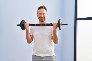 Spor merkezinde bar ve ağırlık diski kullanarak kendinden emin gülümseyen beyaz bir genç.