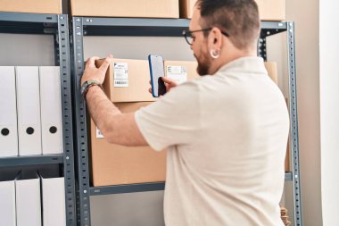 Young hispanic man e-commerce business worker scanning package by smartphone at office