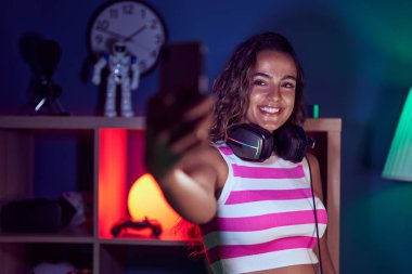 Young beautiful hispanic woman streamer smiling confident make selfie by smartphone at gaming room