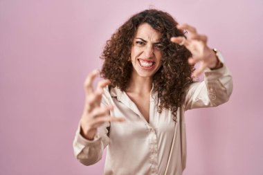 Hispanic woman with curly hair standing over pink background shouting frustrated with rage, hands trying to strangle, yelling mad 