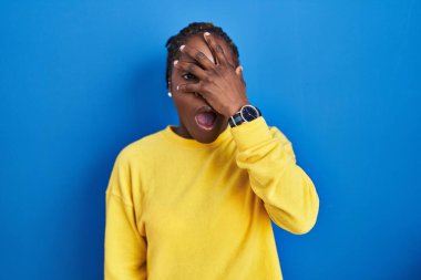 Güzel siyah kadın mavi arka planda dikiliyor. Şoka giriyor. Yüzü ve gözleri kapanıyor, utangaç ifadesiyle parmaklarının arasından bakıyor.. 