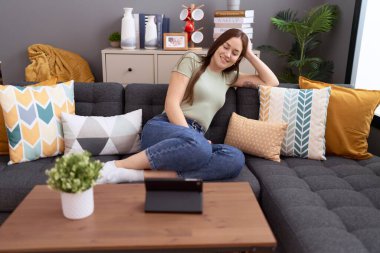 Young beautiful woman watching video on touchpad sitting on sofa at home