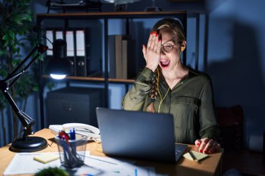 Young blonde woman working at the office at night covering one eye with hand, confident smile on face and surprise emotion. 