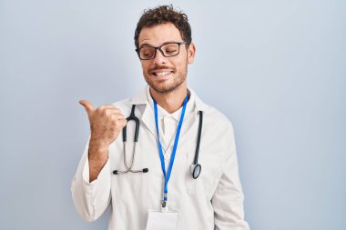 Doktor üniforması ve steteskop giyen genç İspanyol adam gülen yüzle gülümsüyor ve başparmağıyla yan tarafı işaret ediyor.. 
