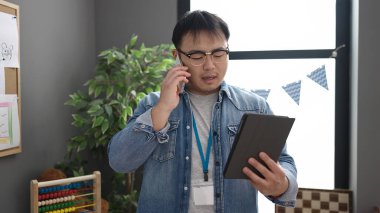 Young chinese man preschool teacher using touchpad talking on smartphone at kindergarten