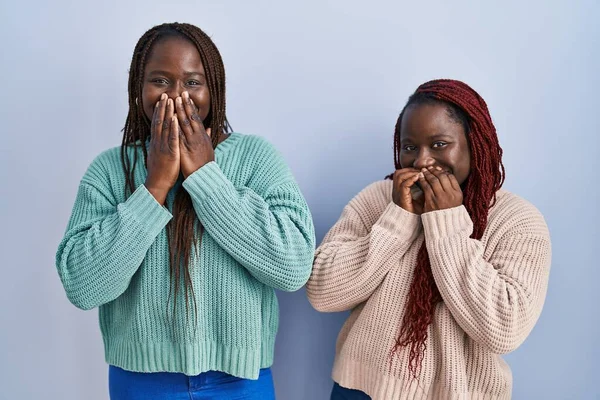 Two african woman standing over blue background laughing and embarrassed giggle covering mouth with hands, gossip and scandal concept