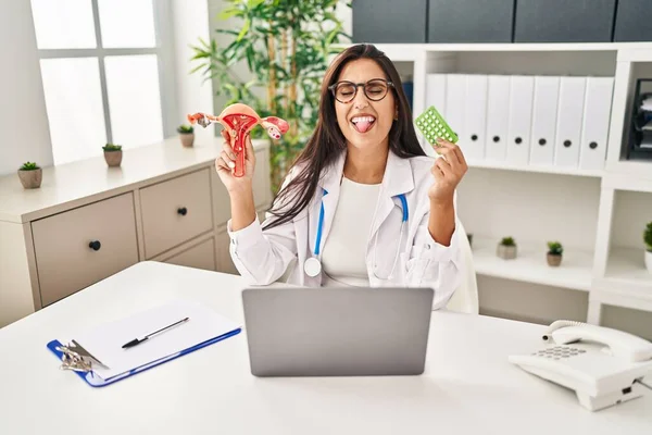 Young hispanic doctor woman holding anatomical female genital organ and birth control pills sticking tongue out happy with funny expression.