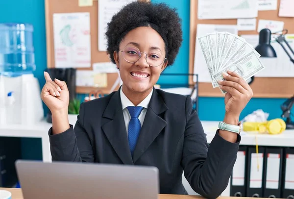 Beautiful african woman with curly hair holding money at the office smiling happy pointing with hand and finger to the side