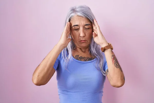 Middle age woman with tattoos standing over pink background with hand on head, headache because stress. suffering migraine.