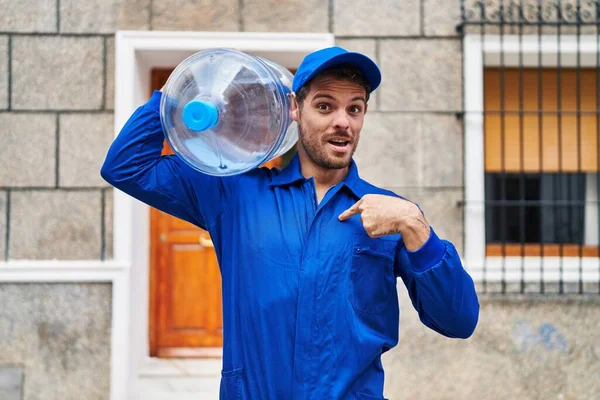 Young hispanic man holding a gallon bottle of water for delivery pointing finger to one self smiling happy and proud