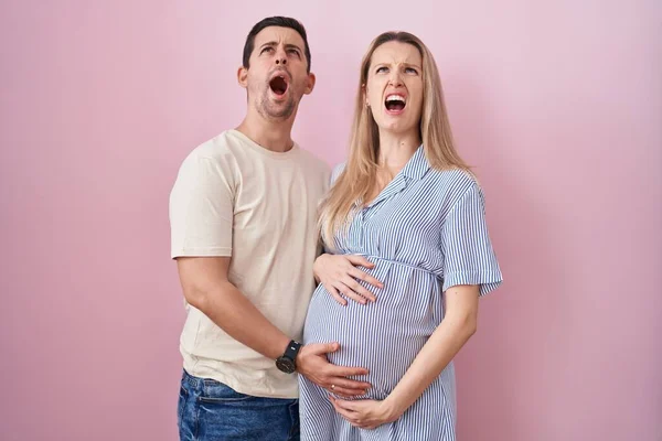 赤ん坊がピンクの背景に立っていることを期待している若いカップルは怒って怒っていると怒っている叫び 怒りで叫んでいる 怒りと攻撃的な考え方 — ストック写真