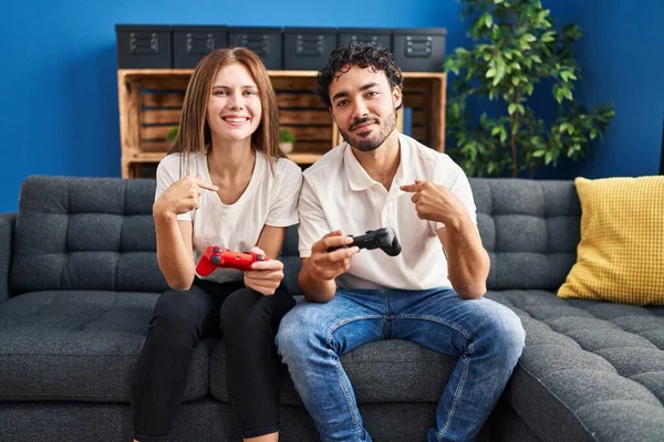 Young couple playing video game holding controller at home pointing finger to one self smiling happy and proud
