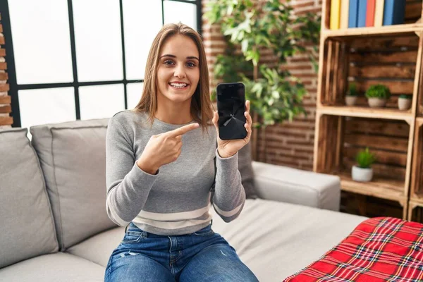 Young woman holding broken smartphone showing cracked screen smiling happy pointing with hand and finger