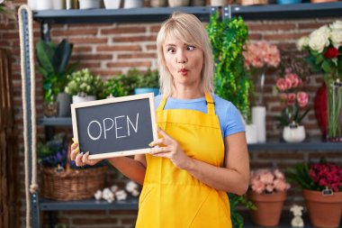 Young caucasian woman working at florist holding open sign making fish face with mouth and squinting eyes, crazy and comical. 
