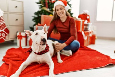 Beyaz bir kadın gülerek evde köpekle oturuyor. Noel ağacı yanında.