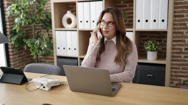 Young beautiful hispanic woman business worker talking on telephone using laptop at office