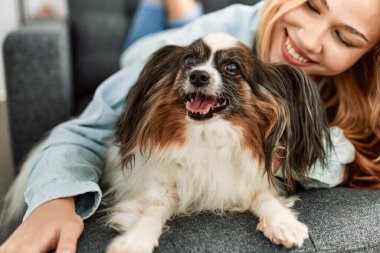 Beyaz bir kadın, evinde köpekle kanepede uzanmış kendinden emin bir şekilde gülümsüyor.