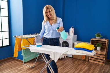 Middle age blonde woman smiling confident ironing clothes at laundry room