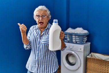 Yaşlı kadın, elinde deterjan şişesiyle baş parmağını kaldırmış, ağzı açık gülümsüyor. 