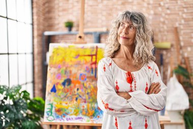Middle age woman artist standing with arms crossed gesture at art studio