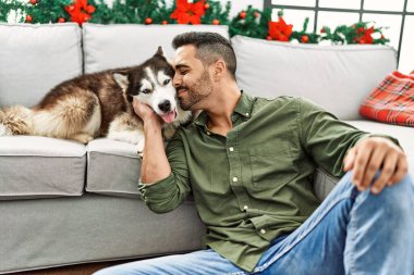 Young hispanic man kissing dog sitting on sofa by christmas decor at home
